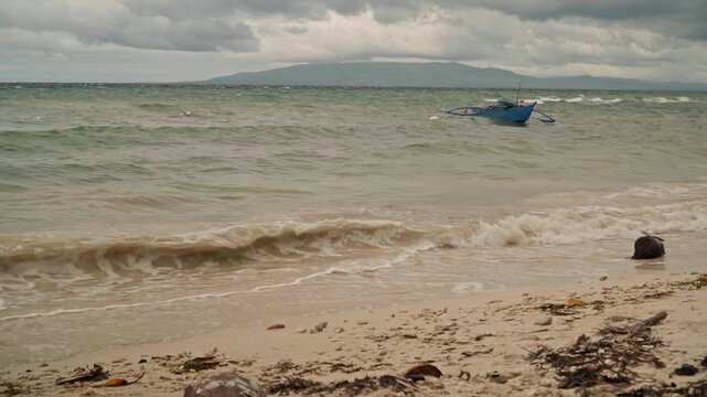 Cloudy beach with fishing boat offshore, seaweed and driftwood scattered on shore, choppy green water under heavy sky, distant island silhouette and moody coastal atmosphere ready for cinematic usage