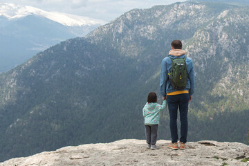 Fototapeta premium Father with backpack holding little daughter hand standing on rocky cliff edge overlooking vast mountain valley with forest and snow capped peaks under cloudy sky, family travel bonding , copy space