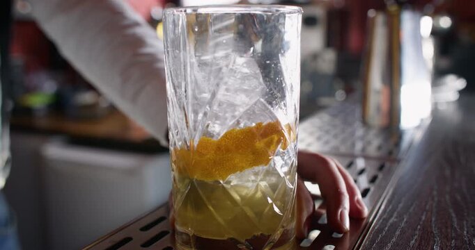 Cocktail making. Drink mixing. Unrecognizable professional male barman stirring ice cubes lemon spiral zest with spoon in transparent glass on bar counter.