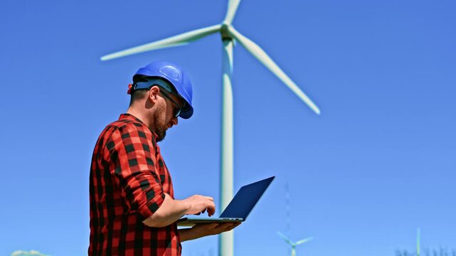 A windmill engineer stands on a wind farm holding a laptop. The engineer looks up at a large wind turbine, focused on their task.