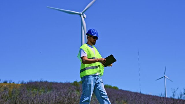 A windmill engineer walks on the ground at a wind farm. He wears a safety vest and hard hat while checking equipment. The wind turbines turn in the background on a clear day.