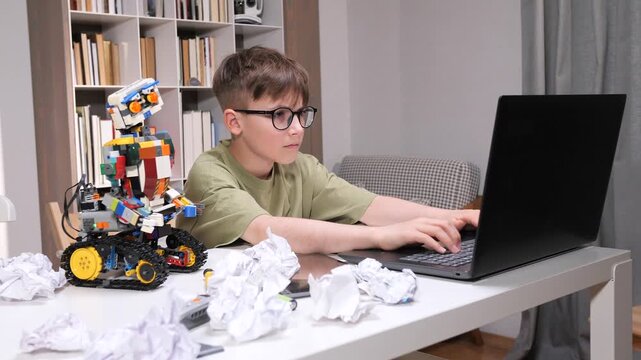 Boy programming a self built robot with building bricks, actively using a laptop on a desk filled with crumpled papers, exploring stem education and technology