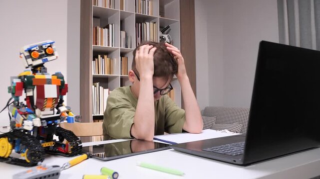 Young boy with glasses holding head in hands, experiencing stress and frustration while sitting at a desk with a robot, laptop, and tablet, engaging in stem education and home learning challenges