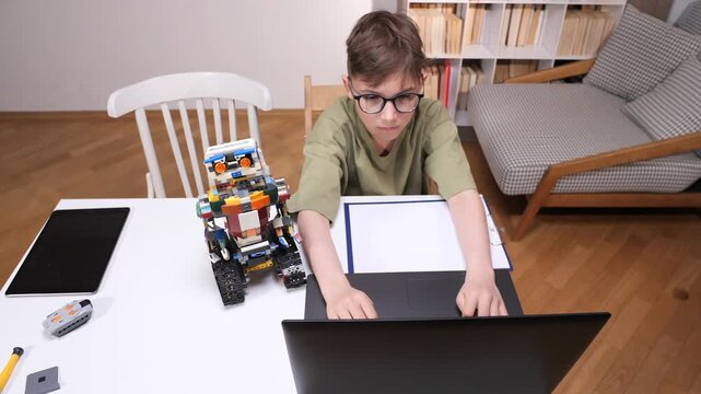 Young boy wearing eyeglasses sitting at a white table, programming a multicolored robot toy with a laptop during a stem education and technology lesson
