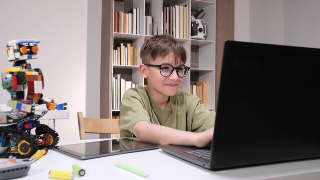 Young male student wearing glasses building and programming a robot with building blocks, using a laptop, showing interest in science, technology, engineering, and mathematics activities
