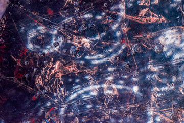Glossy abstract texture shows scratched plastic, water droplets, reflections, and dark blue red...
