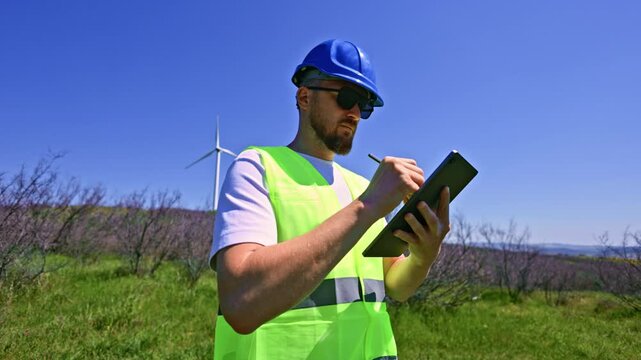 A windmill engineer stands in a green field, wearing a hard hat and safety gear. He is taking notes on a tablet while looking at a nearby wind turbine in clear weather.