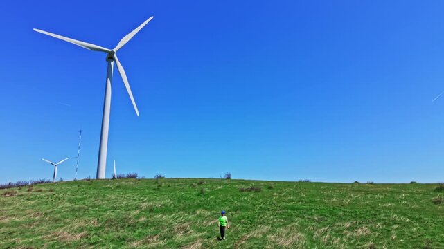 An engineer inspects windmills on a grassy hill. The sky is clear, and the windmills stand tall. The engineer wears a safety vest and looks at the equipment, ensuring it works properly.