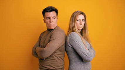 Fototapeta premium Couple standing back to back against a yellow background, expressing frustration and disagreement with arms crossed