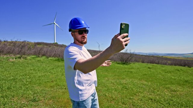 A windmill engineer stands in a green field at a wind farm, wearing a blue hard hat and sunglasses. He is taking a selfie with his phone on a clear sunny day.