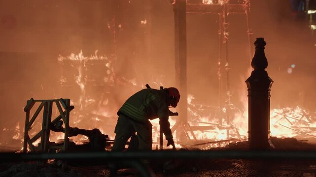 A firefighter in protective gear works at close range among glowing burning falla debris and structural remains during Las Fallas La Crema in Valencia