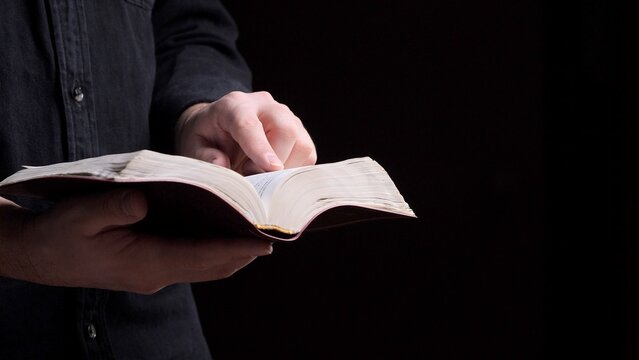 Person's hands holding an open book, likely a bible, with a finger pointing at the text and pages, symbolizing study, faith, devotion, and spiritual guidance against a dark background
