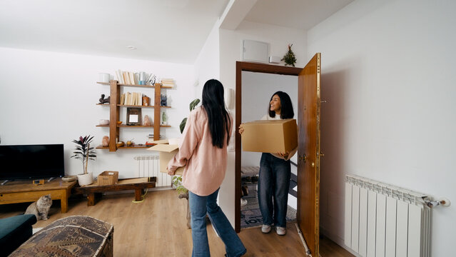 Young women carrying cardboard boxes, relocating to their first new home together, starting fresh in a modern living room