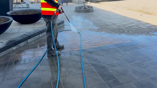 Worker in safety vest using pressure washer to clean paving stones, removing dirt and grime from outdoor walkway on a sunny day