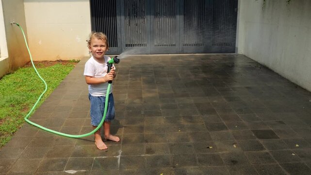 Young toddler playing with garden hose, holding hand sprinkler and playfully spraying water upward and toward camera, creating small rainbow in mist. Little boy enjoys sunny summer day game