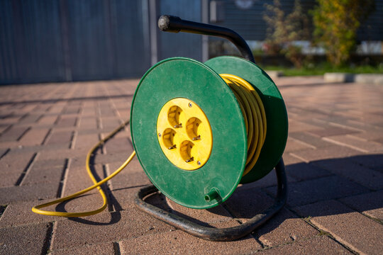 Green cable reel with yellow extension cord on paved courtyard, outdoor power supply for garden equipment in daylight.