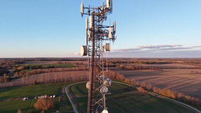 A smooth aerial drone descent alongside a telecommunications tower at golden hour, revealing multiple antenna arrays, microwave dishes, and vast agricultural fields