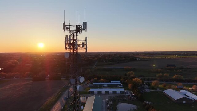 A cinematic 360-degree aerial drone orbit around a 5G cellular tower at golden hour, revealing rural farmland, agricultural buildings, and a vast horizon during a vibrant sunset