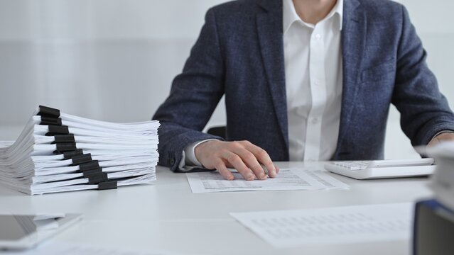 Businessman sorting through stacks of documents on a white desk beside a tall pile of bound papers, symbolizing meticulous financial record keeping and bureaucracy. Audit and taxes