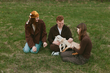 Naklejka premium Three friends outdoors share a gentle moment with a goat kid, sitting on grass, smiling, petting, sunlit scene, casual clothing, peaceful rural vibe, natural colors, candid lifestyle