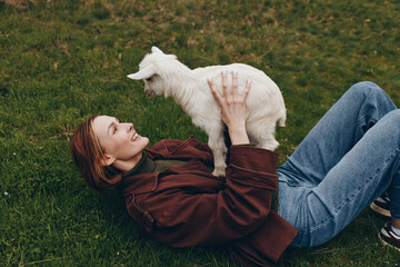 Naklejka premium Young woman lying on grass holds a playful white baby goat above her while smiling, enjoying a relaxed outdoor moment in casual clothing during a lifestyle photoshoot.