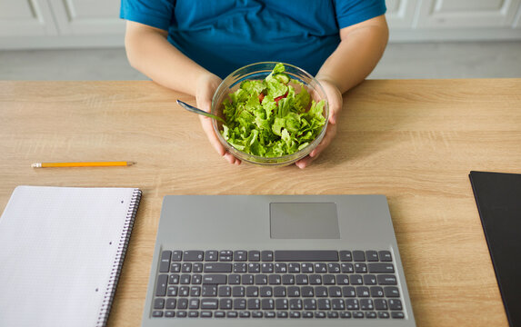 Diet salad at home by laptop. A plus-size overweight man eats greens, checking tasks in the kitchen and balancing remote work. Mindful eating supports healthy weight loss. Health and nutrition.