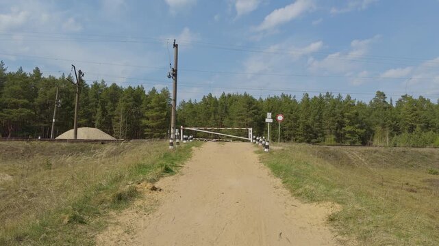 Secluded pine path with barrier. Untrodden gravel track bordered by aged posts and trees. Isolated country lane with weathered barrier amidst dense pine forest under drifting clouds