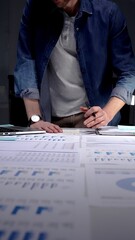 Entrepreneur standing at a desk in a dark office, studying financial reports and charts by lamp...