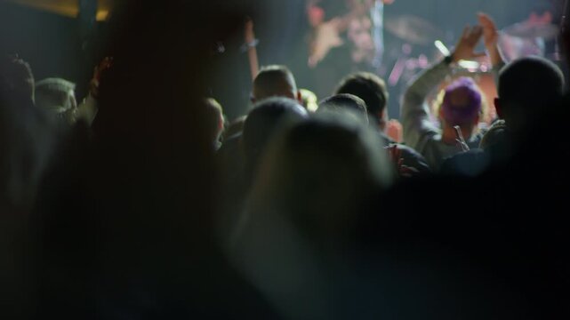Wide frame of concert crowd viewed from behind, multiple hands and arms raised, teal green bokeh stage light illuminates venue from front