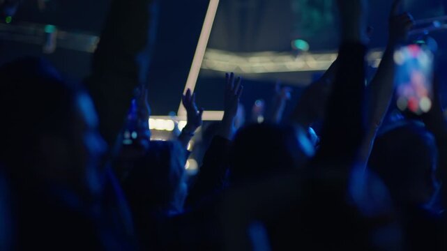 Audience members raise arms and hands at concert, white illuminated truss and strip lighting above, teal blue stage atmosphere from front