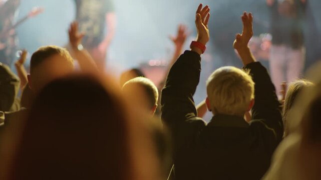 Young fan raises hand with red wristband amid crowd at live music event, warm golden stage light and smoke haze illuminate the scene from behind
