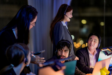 Fototapeta premium Multiethnic corporate colleagues working late. Professional female manager standing and assisting male operator wearing headset in a modern office with blurred foreground.