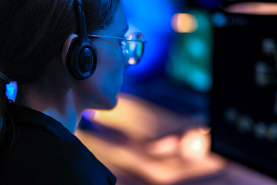 Over the shoulder view of a female IT support agent wearing headset. Professional woman looking at computer screen working late night shift in a glowing dark office.