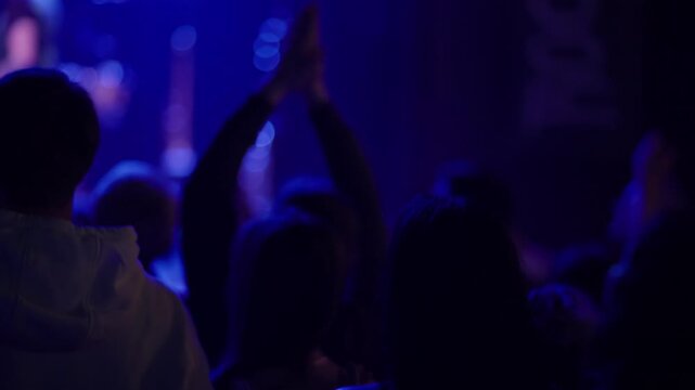 Multiple fans raise and clap hands at live concert, white wristband clearly visible on one raised arm, blue stage light fills background