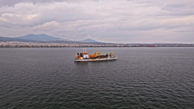 High-quality aerial footage of a dredger support vessel, also known as a subsea cable ship, operating in a busy industrial port. The vessel is designed for underwater cable installation, maintenance, 