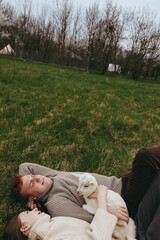 Naklejka premium Relaxed couple outdoors on a grassy field cuddle a white goat kid, cozy sweaters, natural light, intimate lifestyle moment, tranquil rural setting, warm affection and companionship