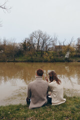 Fototapeta premium Couple sitting by a calm lake in autumn, bundled in knit sweaters, sharing a quiet moment. Warm tones, soft light, and a gentle breeze create a peaceful mood about connection and nature.