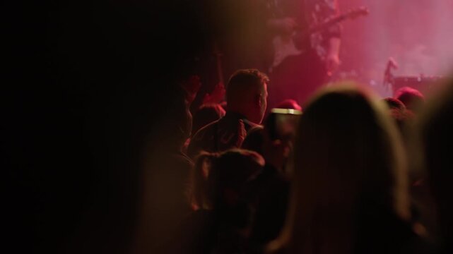 Fan raises arm holding phone above head in warm golden hazy concert atmosphere, other audience members visible raising hands around