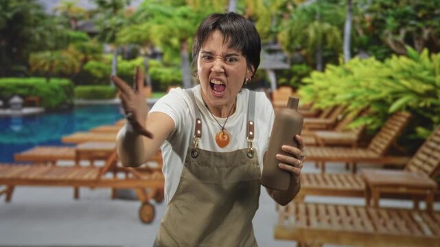 Woman wearing apron and short hair grips pottery bottle and flings wet clay with raised hand in studio; anger craft.