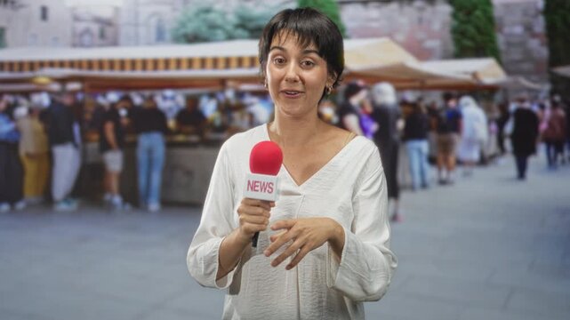 Woman holding red microphone labeled news and smiling, finger to lips hush gesture on a crowded street scene; cheerful reporting.