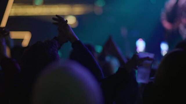 Crowd at live concert raises hands, teal blue stage light and neon strip element visible above, performer silhouette visible in background