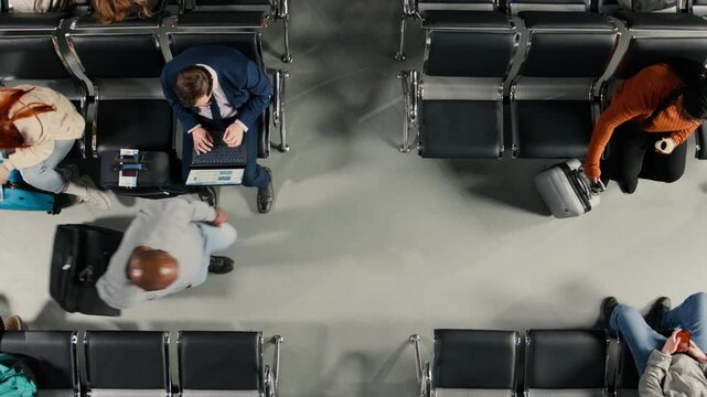 Top down view of travelers rushing through busy airport with people killing time on chairs. Tourists with trolleys navigating hub next to people waiting for plane, high volume passenger flow concept