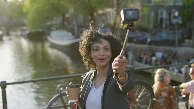 Influencer woman vlogging with a selfie stick and drinking coffee on a bridge over a canal in amsterdam at sunset