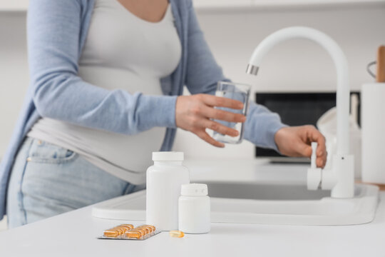 Folic acid pills on counter against pregnant woman pouring tap water in kitchen, closeup
