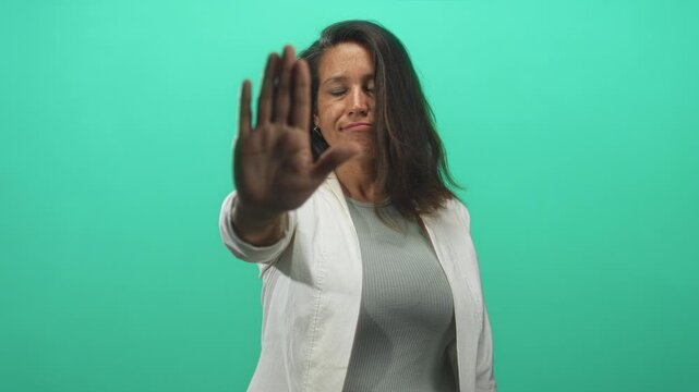 Brunette middle age woman shows open palm stop gesture toward camera in studio green backdrop; boundary setting defiance.