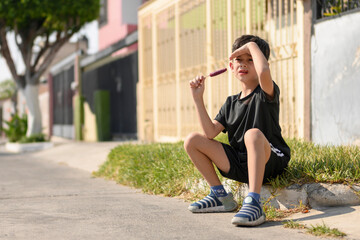 Little boy sitting on a sidewalk eating a popsicle and shading his eyes from the sun, summer in the city. © alejandro
