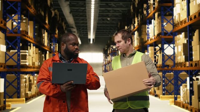 Two warehouse colleagues organizing pallets and packages with laptop, checking inventory control information in industrial depot. Teamwork enhances operational workflow and e-commerce processes.