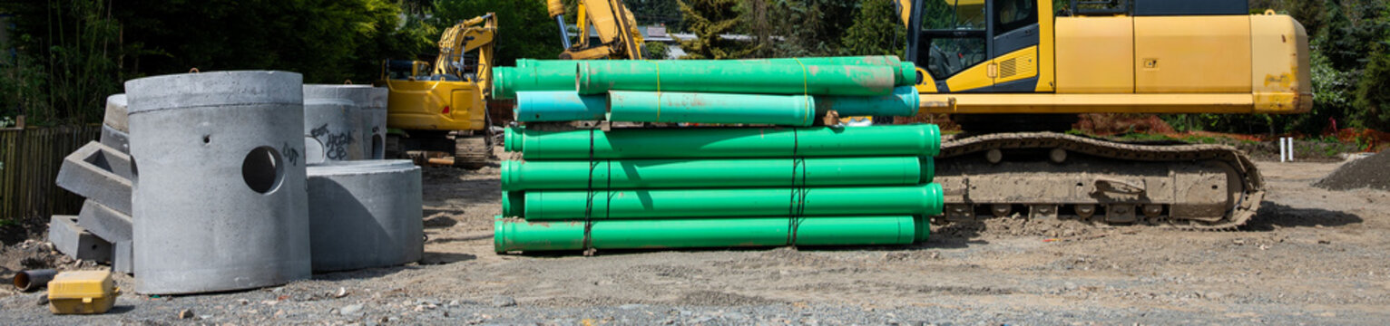 Precast concrete underground utility access vault and stack of 6 inch pipes for stormwater system sitting on construction site ready for installation, excavator in background
