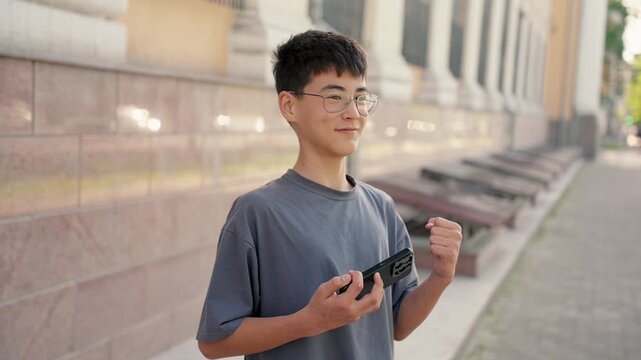 Smiling student in casual attire reacting to good news on smartphone with positive emotions. Real people, authentic success and achievement concept.