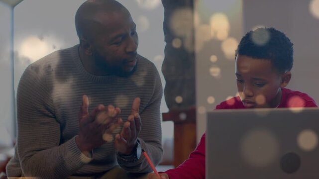 Dad leaning in guiding son writing with animated bokeh floating over laptop, paper for home study